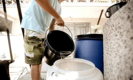 Person pouring water from a black bucket into a white storage container beside a blue barrel